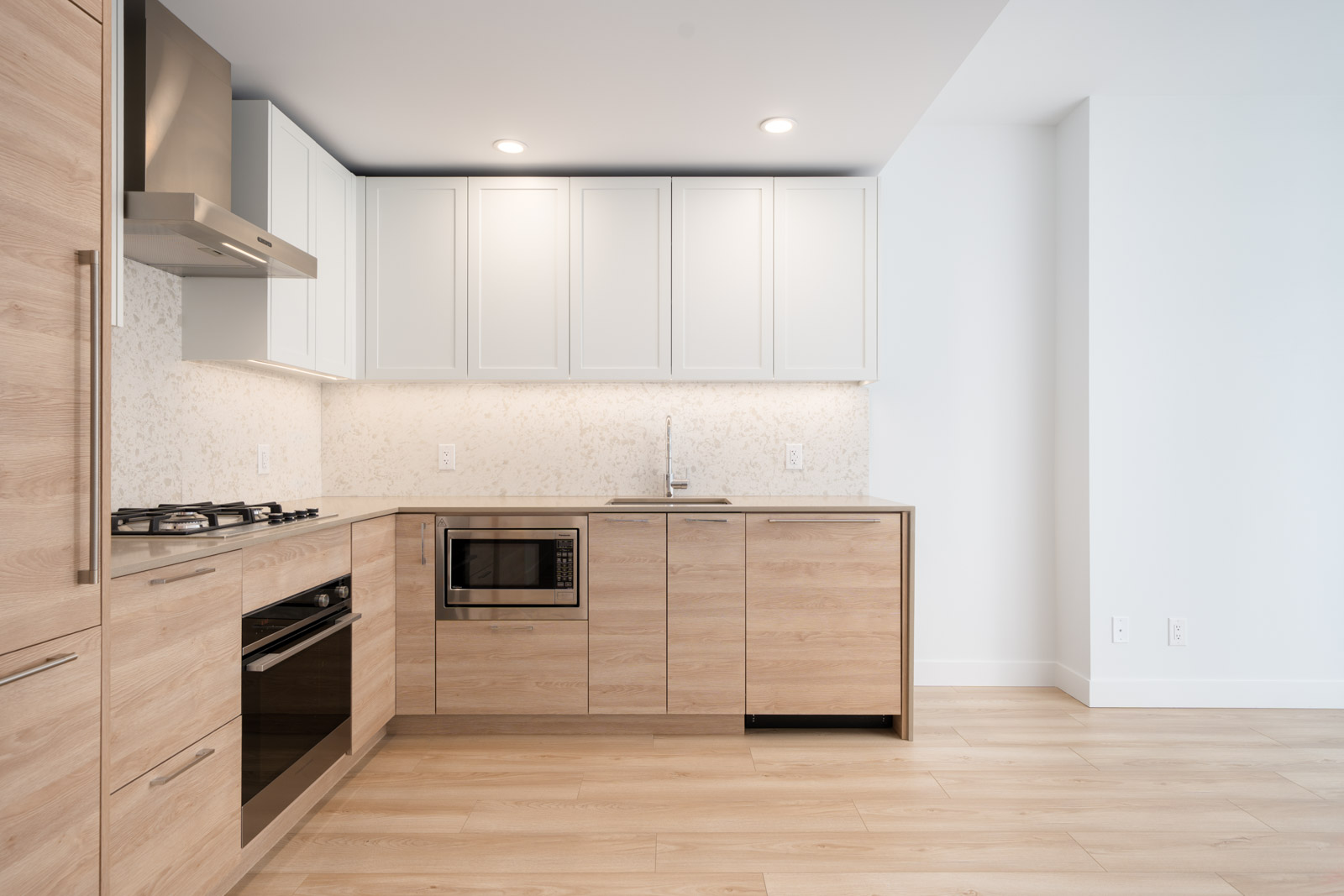 Modern kitchen with light wood cabinets, white upper cabinets, stainless steel appliances, gas stove, built-in oven, microwave, and light-colored flooring.