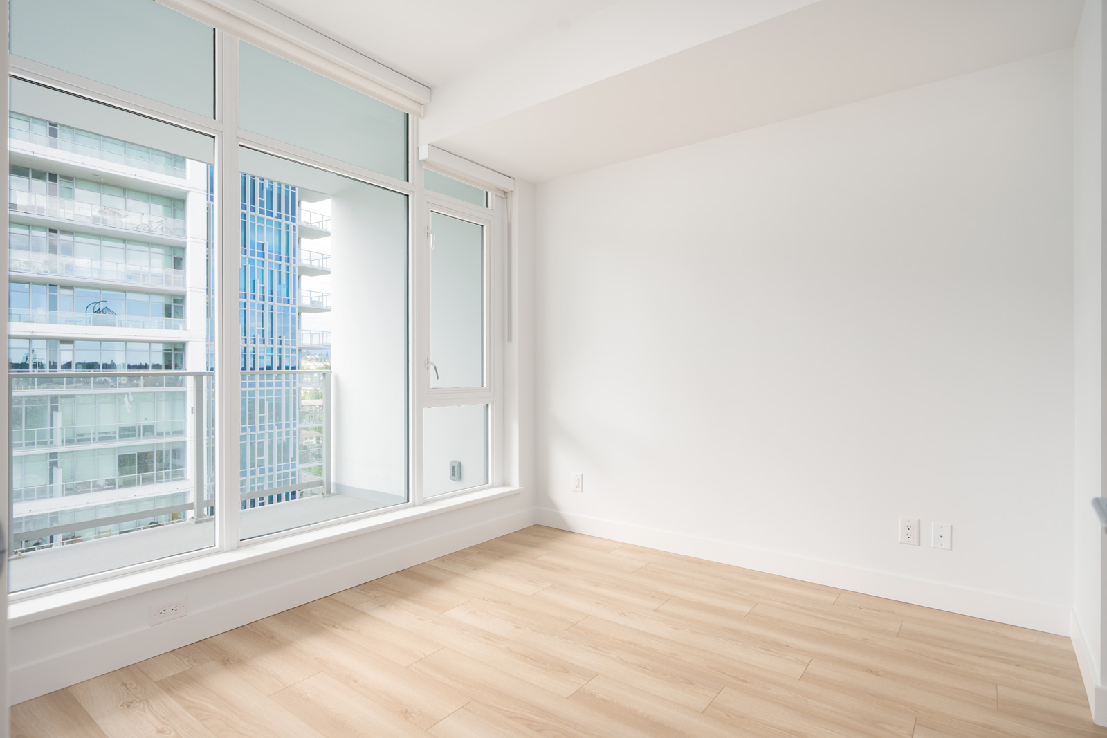 Empty, modern room with light wood flooring, white walls, and large floor-to-ceiling windows overlooking nearby apartment buildings.