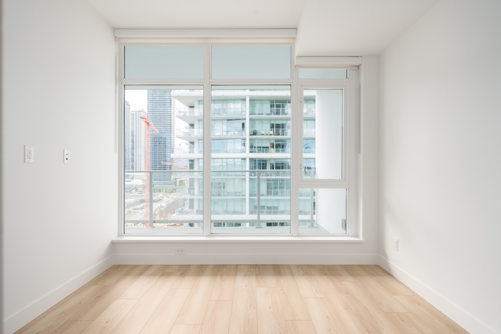 Bright, empty room with light wood flooring, white walls, and large windows overlooking modern high-rise buildings and a construction crane outside.