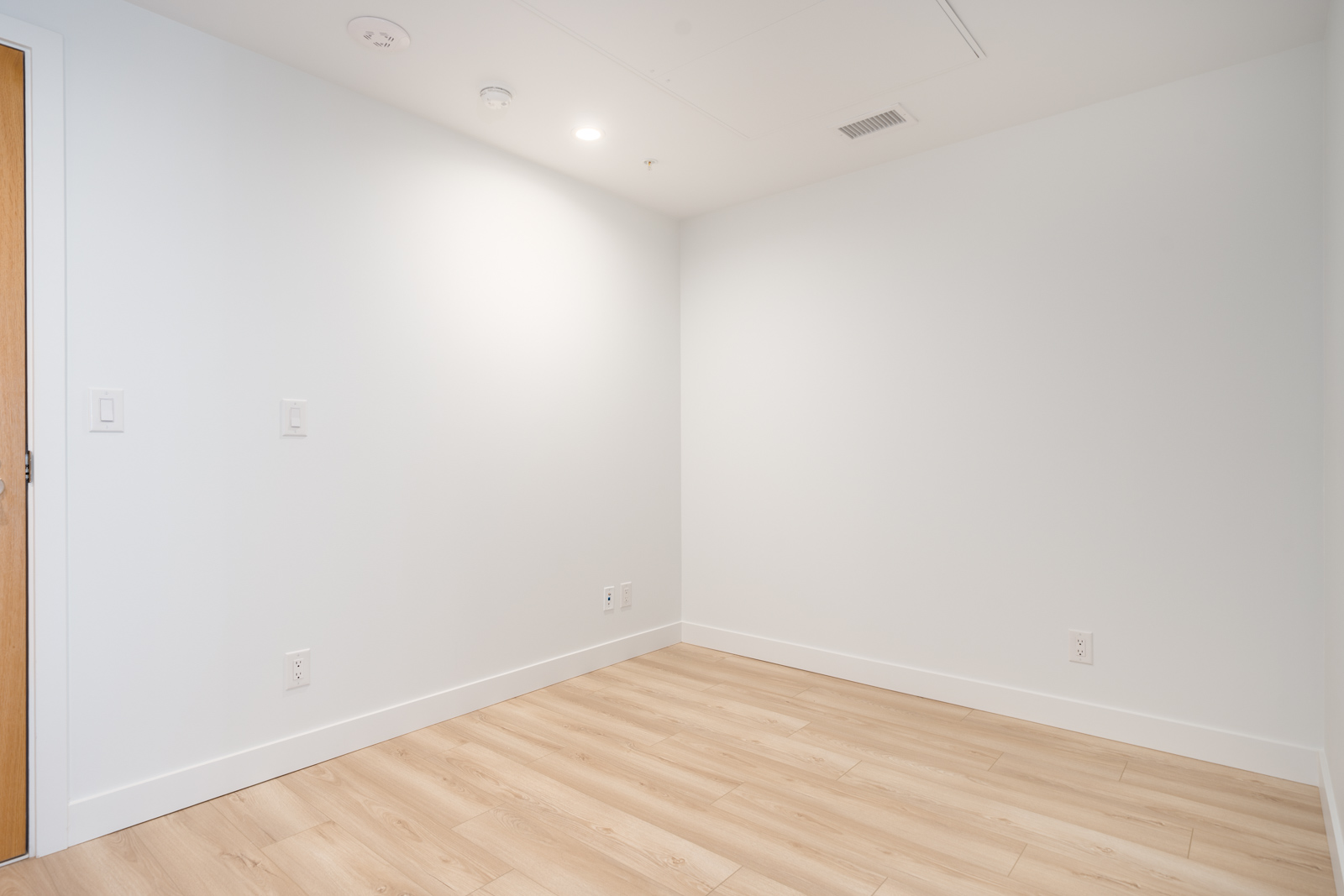Empty room with plain white walls, light wood flooring, and ceiling fixtures, viewed from one corner with a closed wooden door partially visible on the left.