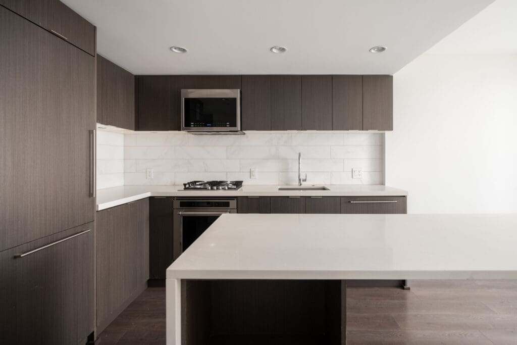 Modern kitchen with dark wood cabinets, white countertops, built-in appliances, and a central island under recessed ceiling lights.