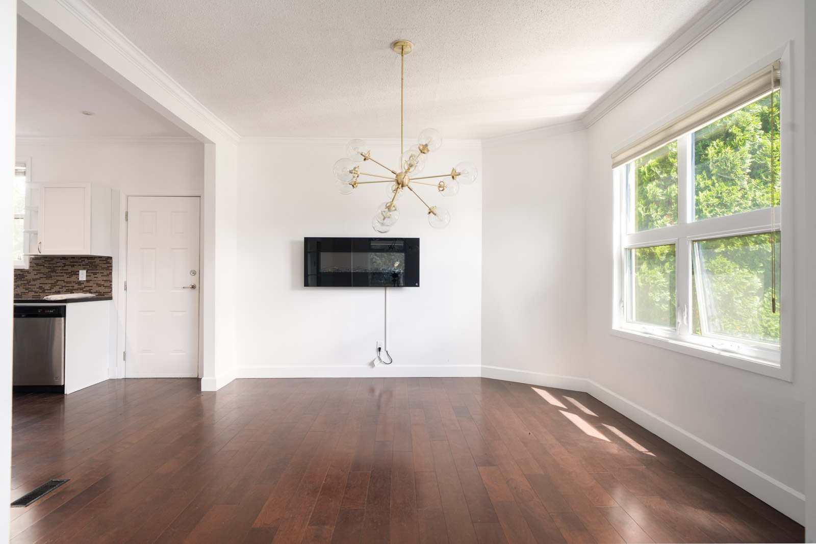 Bright, empty room with white walls, large window, hardwood floor, modern chandelier, and a wall-mounted TV bracket. Kitchen visible through an open doorway.