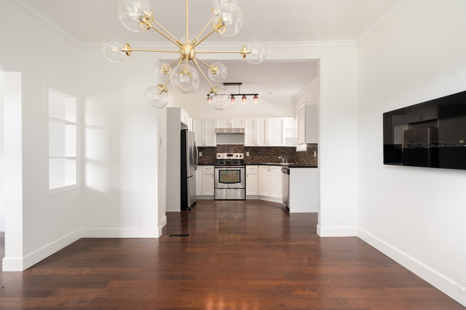 Modern kitchen with stainless steel appliances, white cabinets, dark backsplash, and open layout; hardwood floors and a gold chandelier in the adjacent dining area.