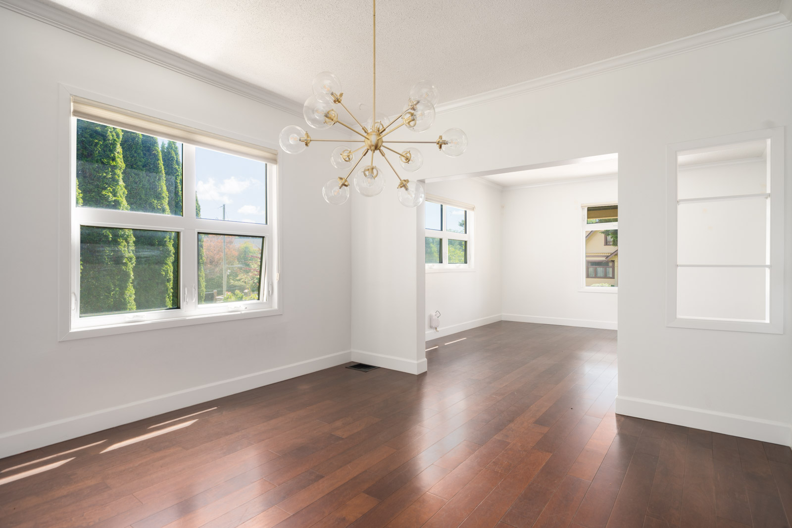 Bright, empty room with large windows, white walls, dark wood floors, and a modern gold chandelier. Natural light fills the space, and a green view is visible outside.