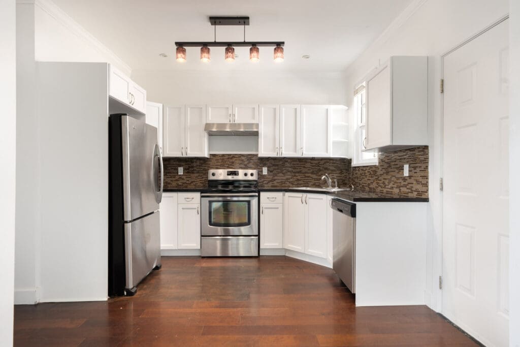 Modern kitchen with white cabinets, stainless steel appliances, brown mosaic backsplash, dark wood flooring, and a rectangular ceiling light fixture.