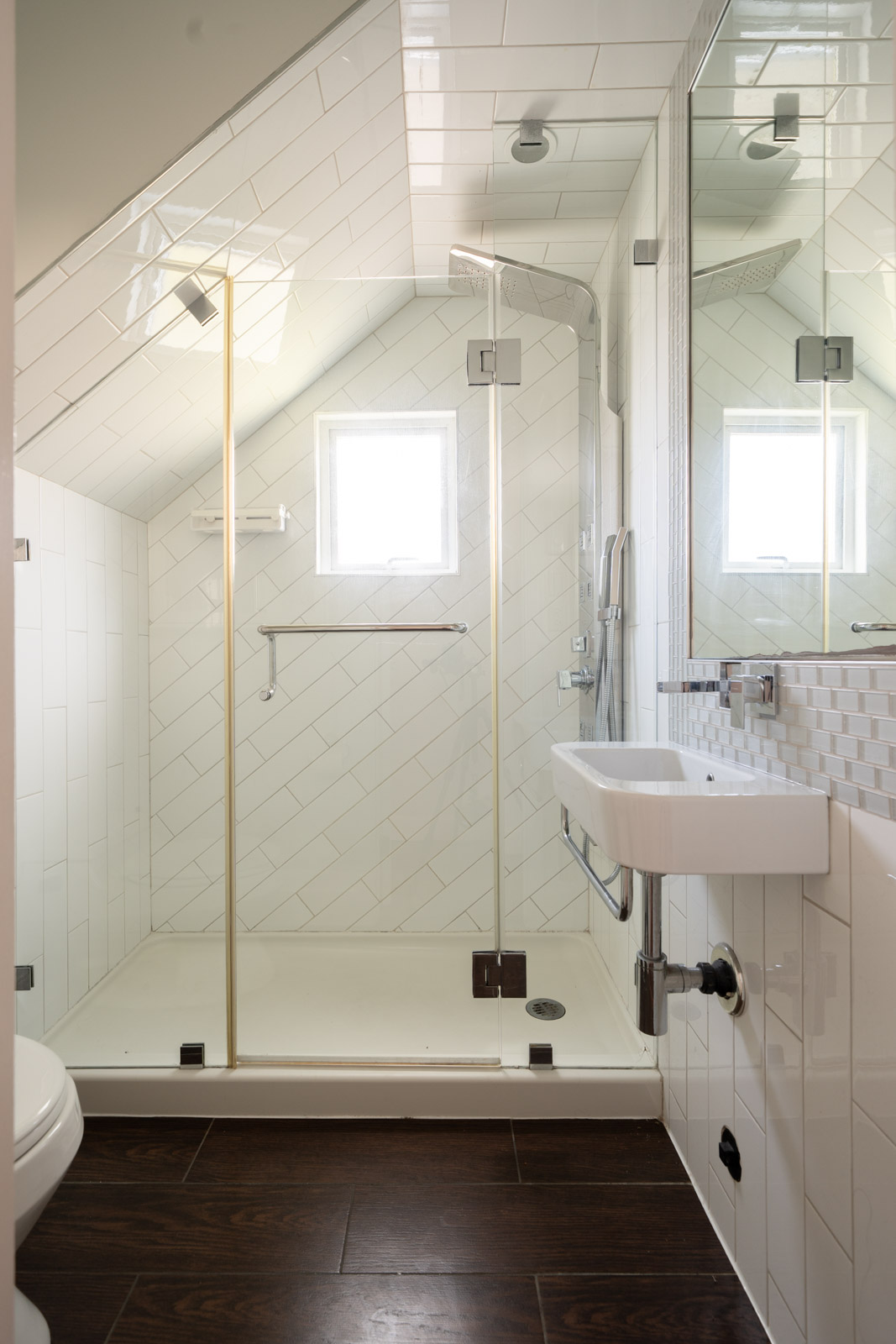 Small, modern bathroom with a glass shower under a sloped ceiling, white tile walls, a wall-mounted sink, and dark wood-look flooring.