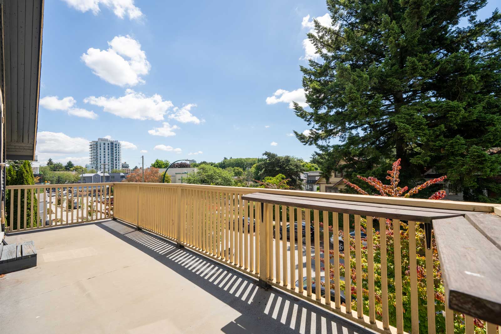 Large outdoor balcony with wooden railing, overlooking trees, buildings, and a distant high-rise under a bright blue sky with scattered clouds.