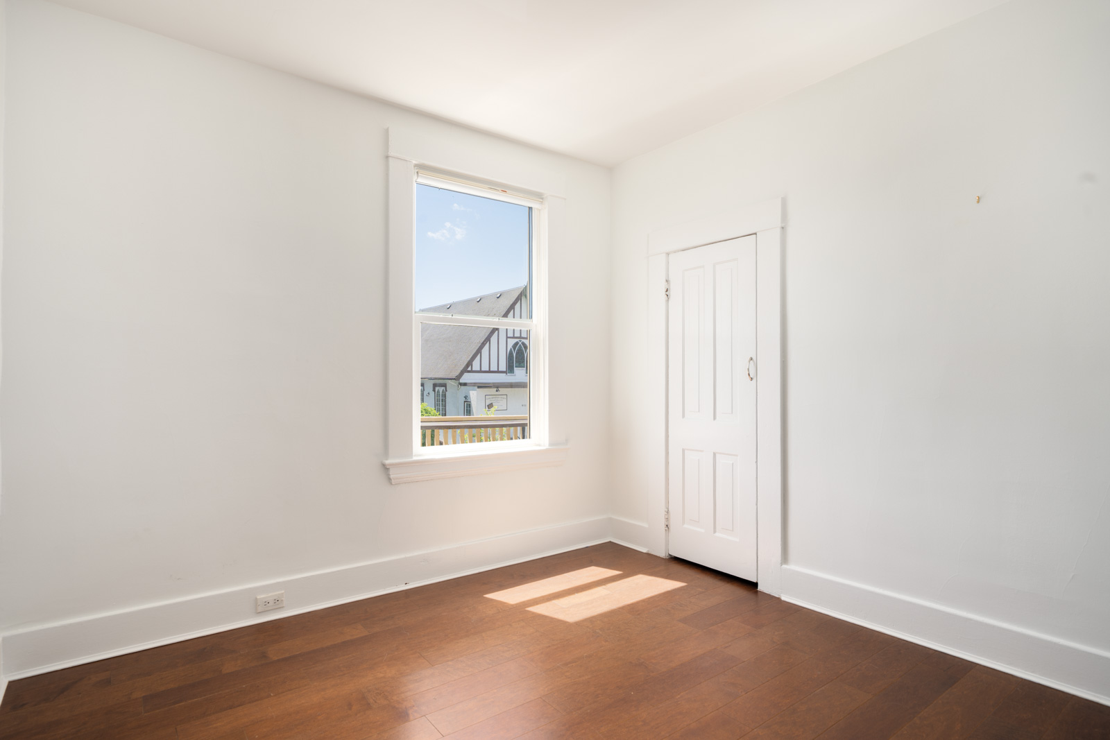 Small empty room with white walls, wooden floor, a window letting in sunlight, and a closed white door. A building is visible outside the window.