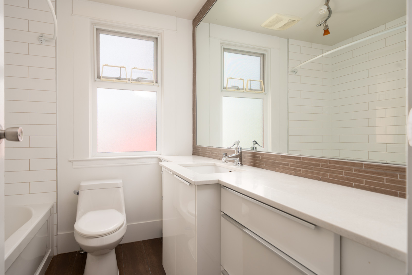 Modern bathroom with white tile walls, a white toilet, white vanity with sink, large mirror, and two frosted windows.