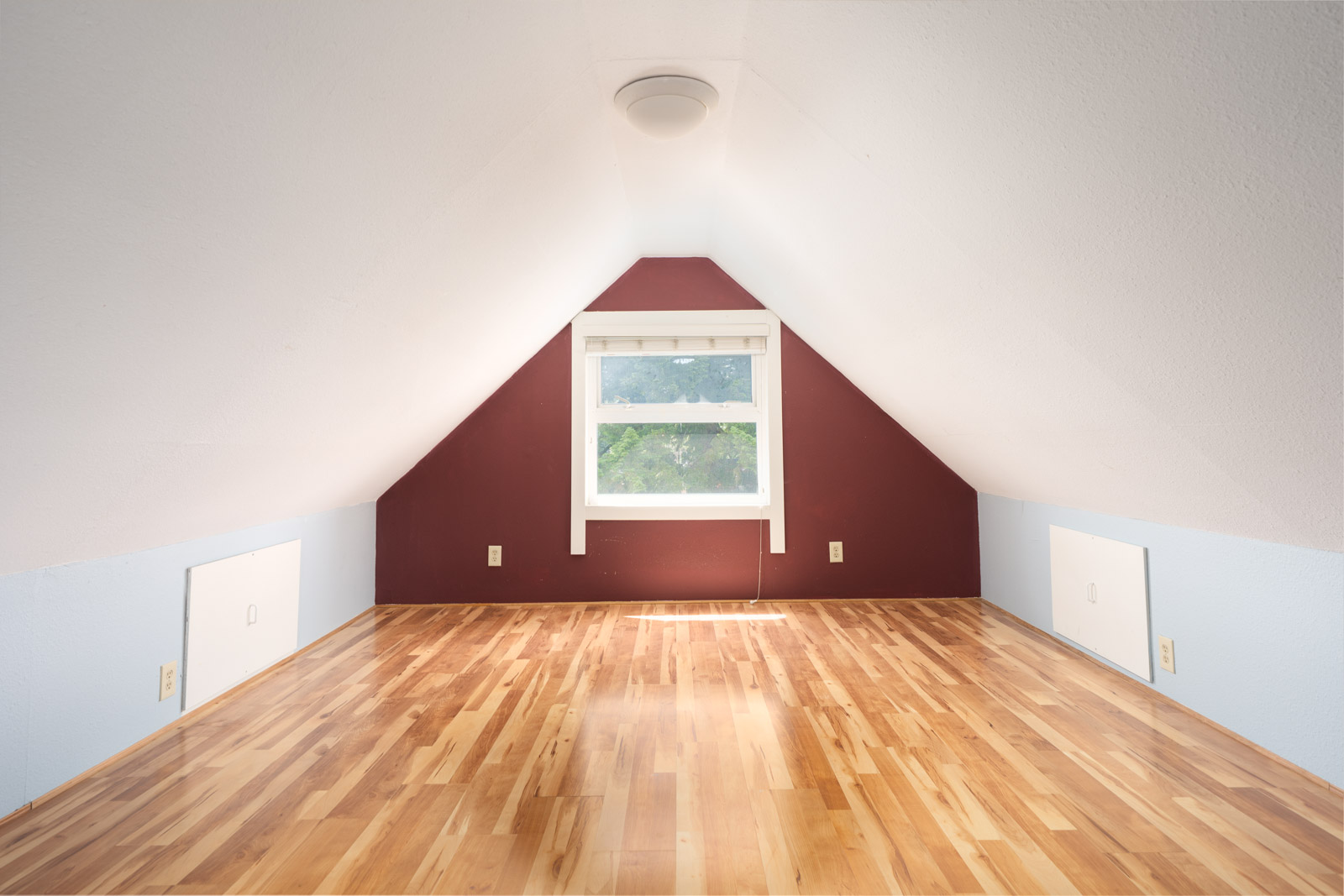 Unfurnished attic room with sloped ceiling, wood floor, maroon accent wall, small window, and light fixture on ceiling.