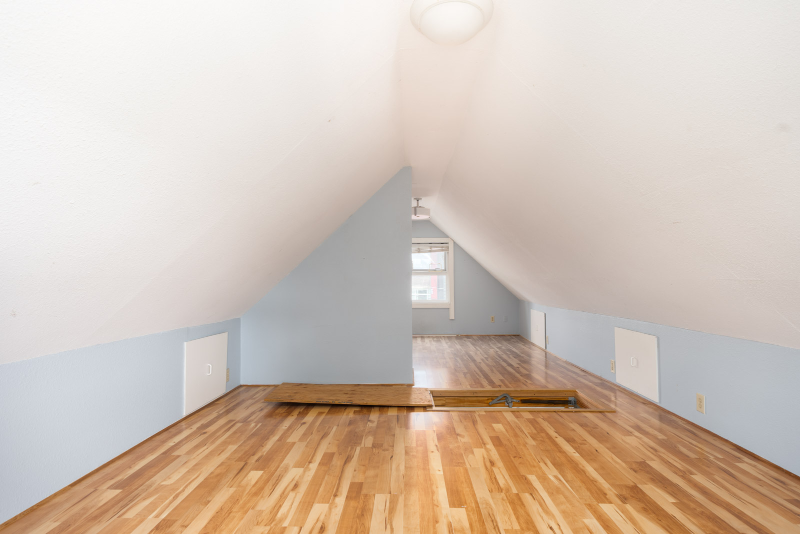 A finished attic room with sloped white ceiling, light blue walls, wood flooring, small storage doors, and an open attic access panel on the floor.