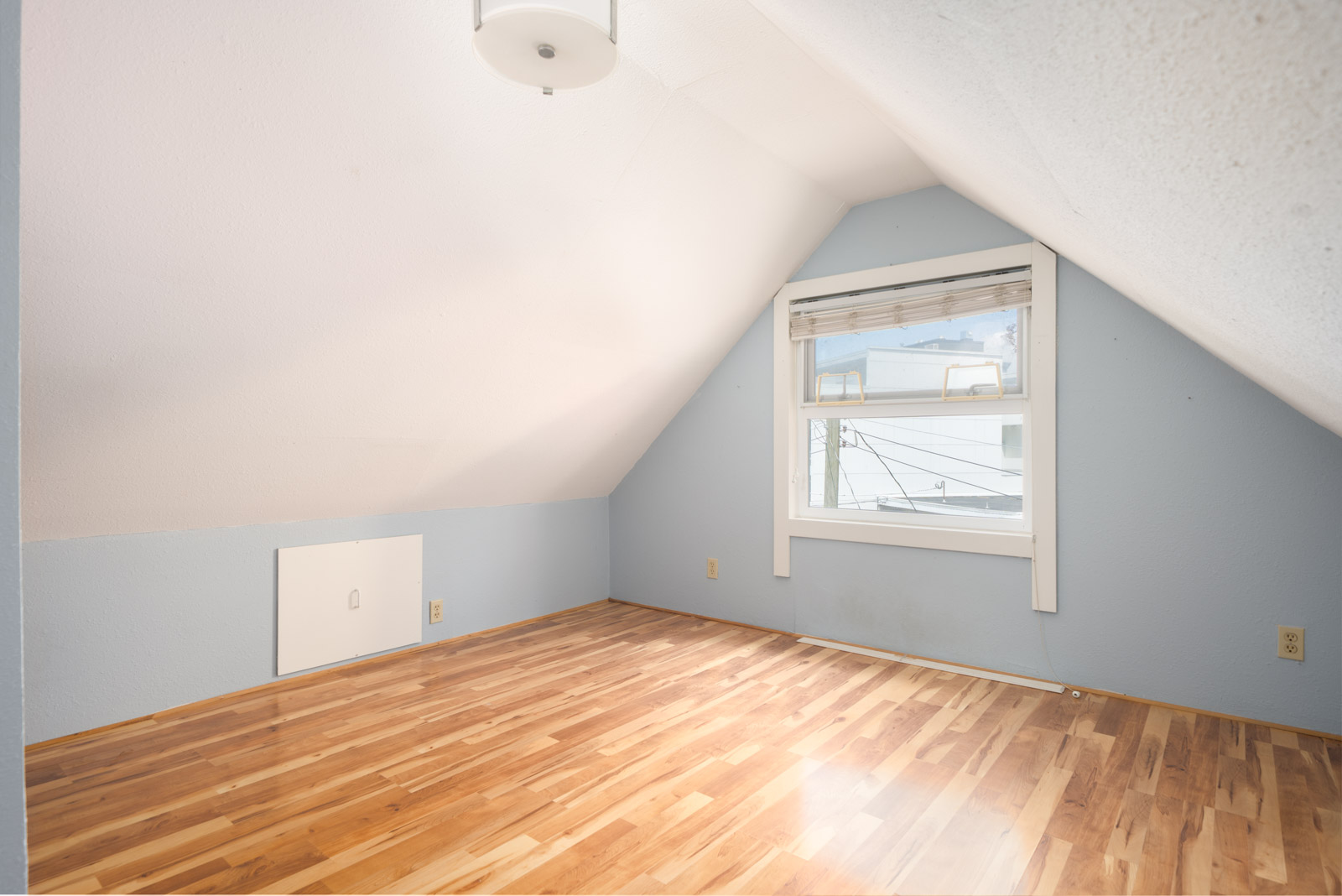 Small attic room with sloped ceiling, light blue walls, a window with blinds, wood flooring, ceiling light fixture, and a small white access door on the left wall.