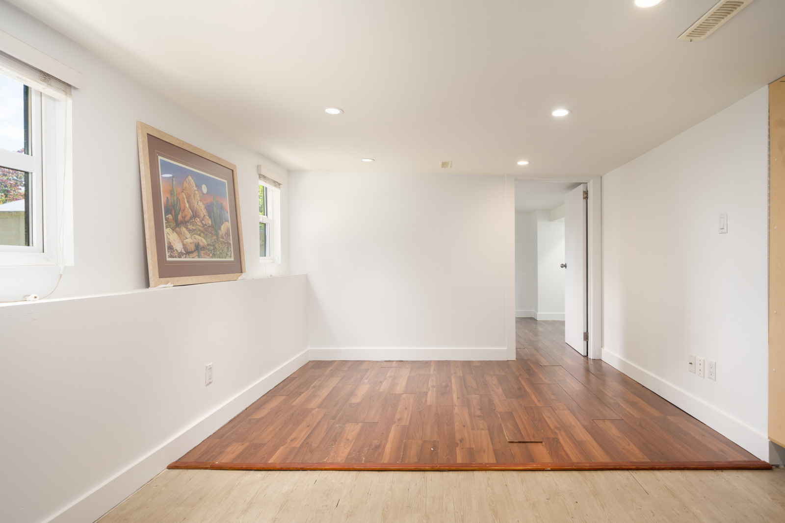 A bright, empty room with white walls, recessed lighting, two windows, framed artwork on a ledge, and a section of laminate wood flooring.