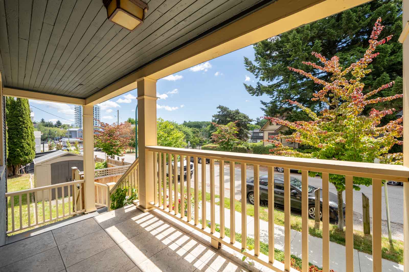 View from a covered front porch with wooden railings, looking out onto a suburban street with cars, trees, and houses under a sunny sky.