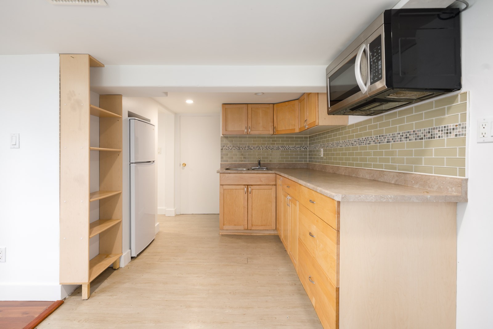 A small kitchen with light wood cabinets, a countertop, a refrigerator, a microwave, open shelving, and a tiled backsplash.