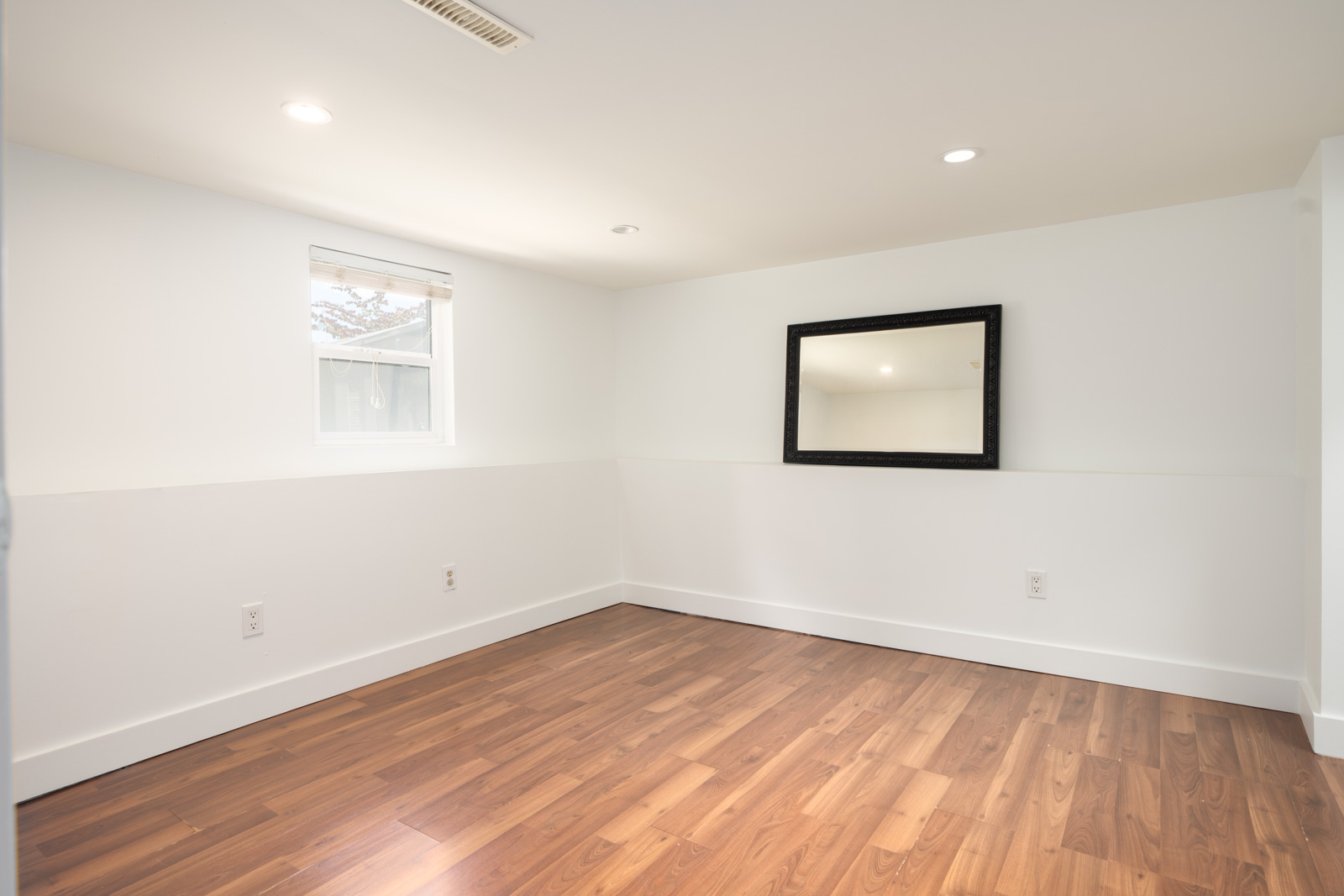 Empty room with white walls, recessed ceiling lights, a small window with blinds, a wall-mounted mirror, and wood flooring.