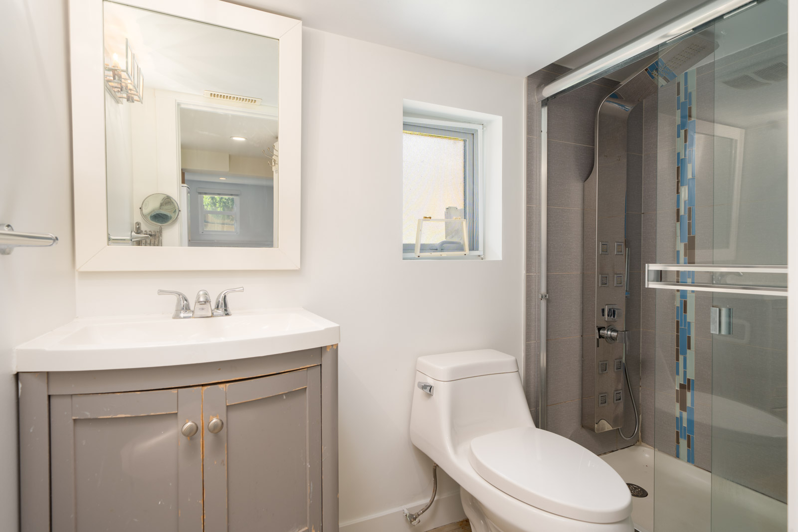 Modern bathroom with a gray vanity, rectangular mirror, white toilet, small window, and a glass-enclosed shower with gray and blue tile accents.