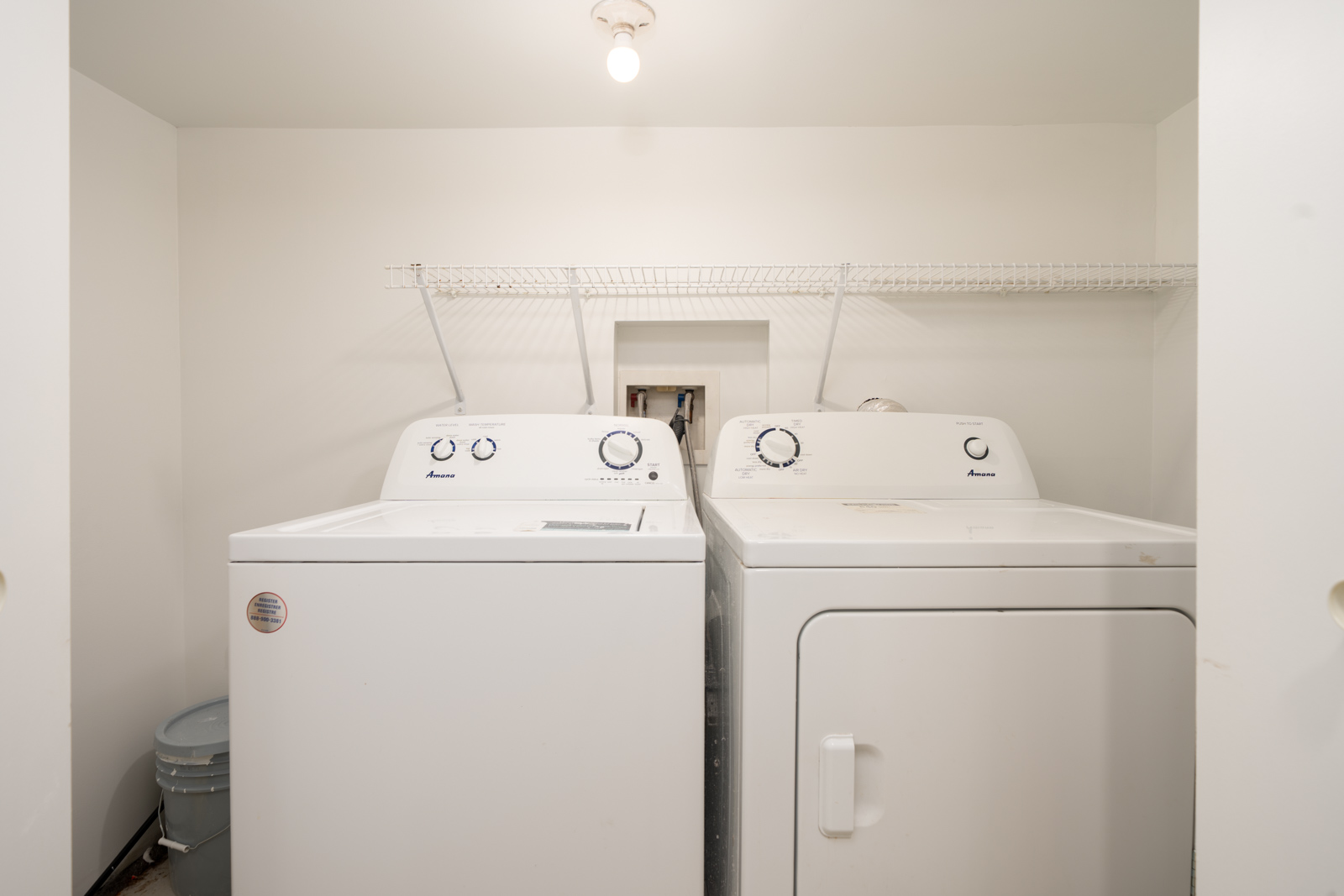 Two white front-loading machines, a washing machine and a dryer, are placed side by side in a small laundry room with a wire shelf above them.
