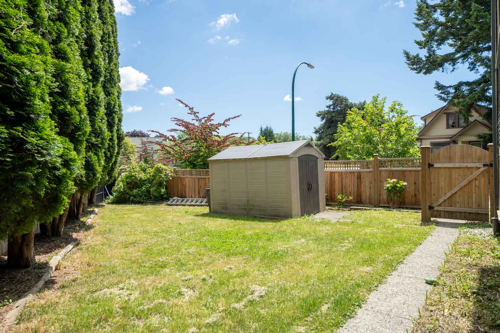 A backyard with a storage shed, green grass, tall trees on the left, a wooden fence, and a paved walkway under a clear blue sky.