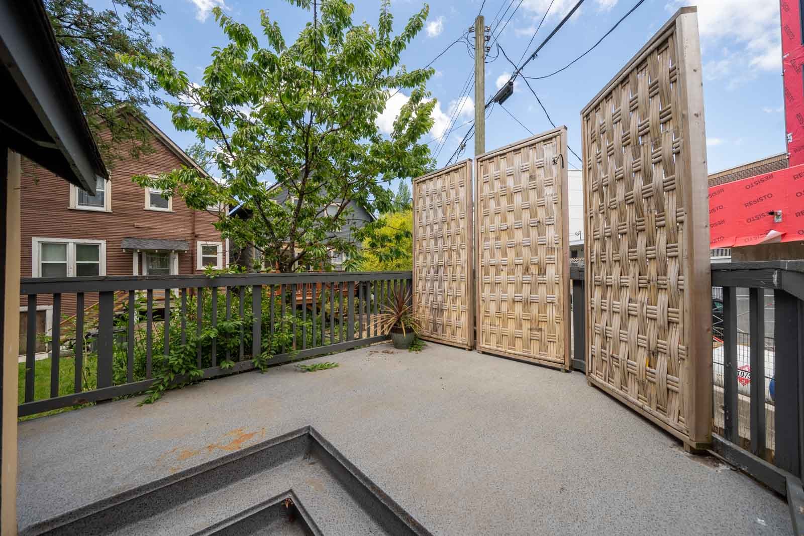 A residential outdoor patio with gray flooring, wooden privacy screens, railing, and a small potted plant; houses and power lines are visible in the background.