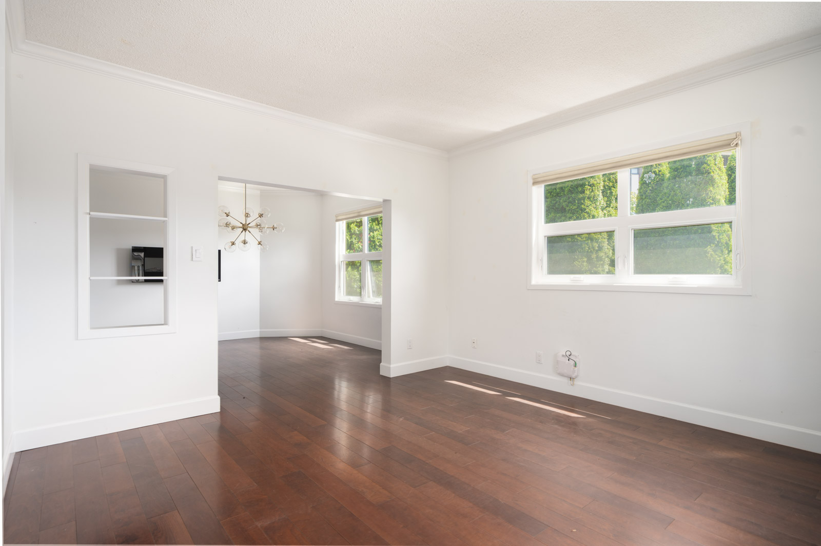 Empty room with white walls, large window, dark wood floors, built-in shelf, and an open doorway leading to another well-lit space with a modern chandelier.