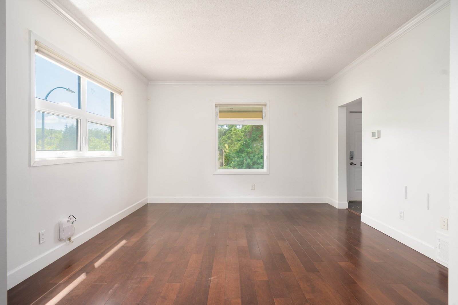 Empty room with white walls, a large window on the left, a smaller window in the center, and dark hardwood floors. A doorway leads to another area on the right.