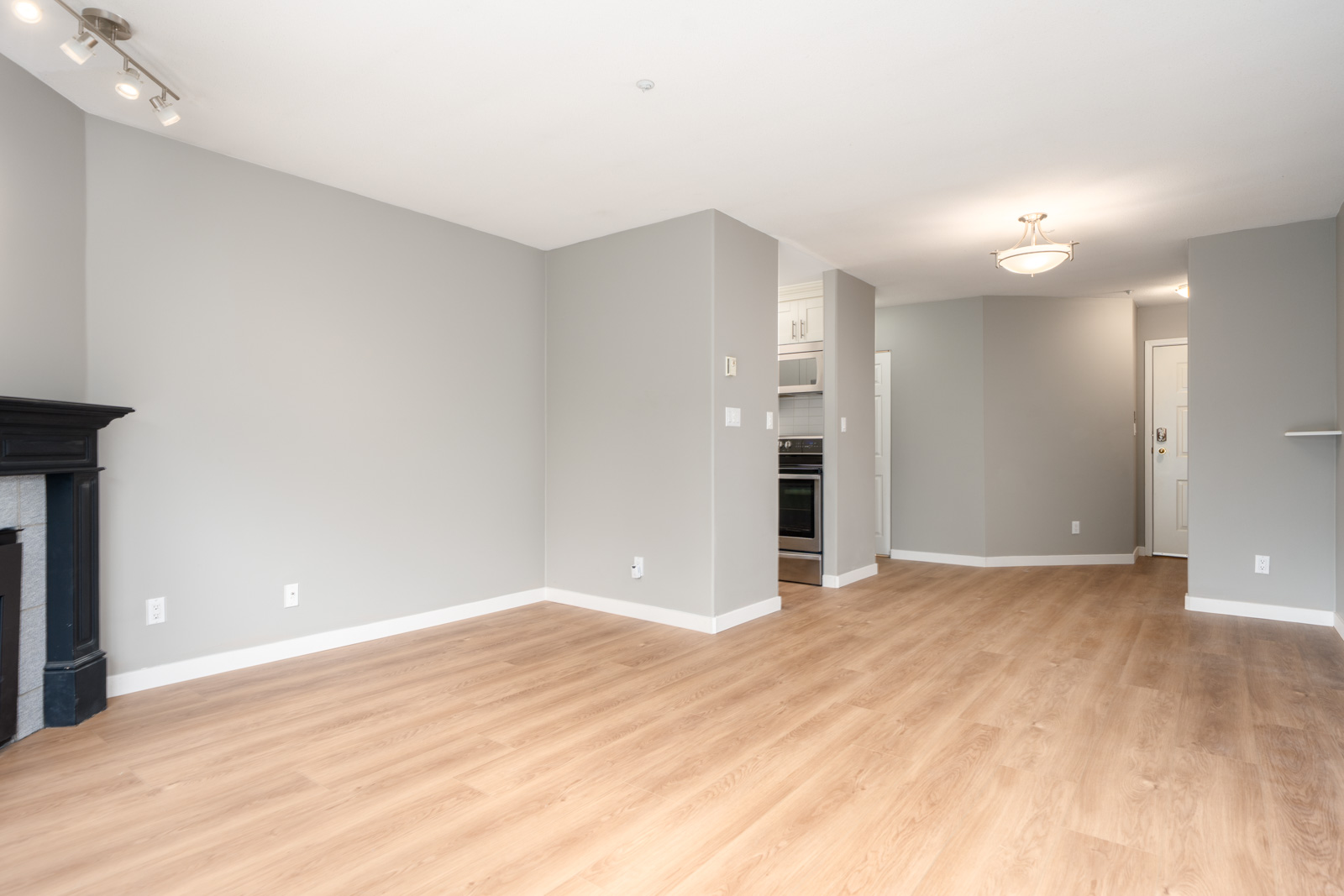 Unfurnished living area with light wood flooring, gray walls, a black fireplace on the left, and an open view to the kitchen and entryway in the background.