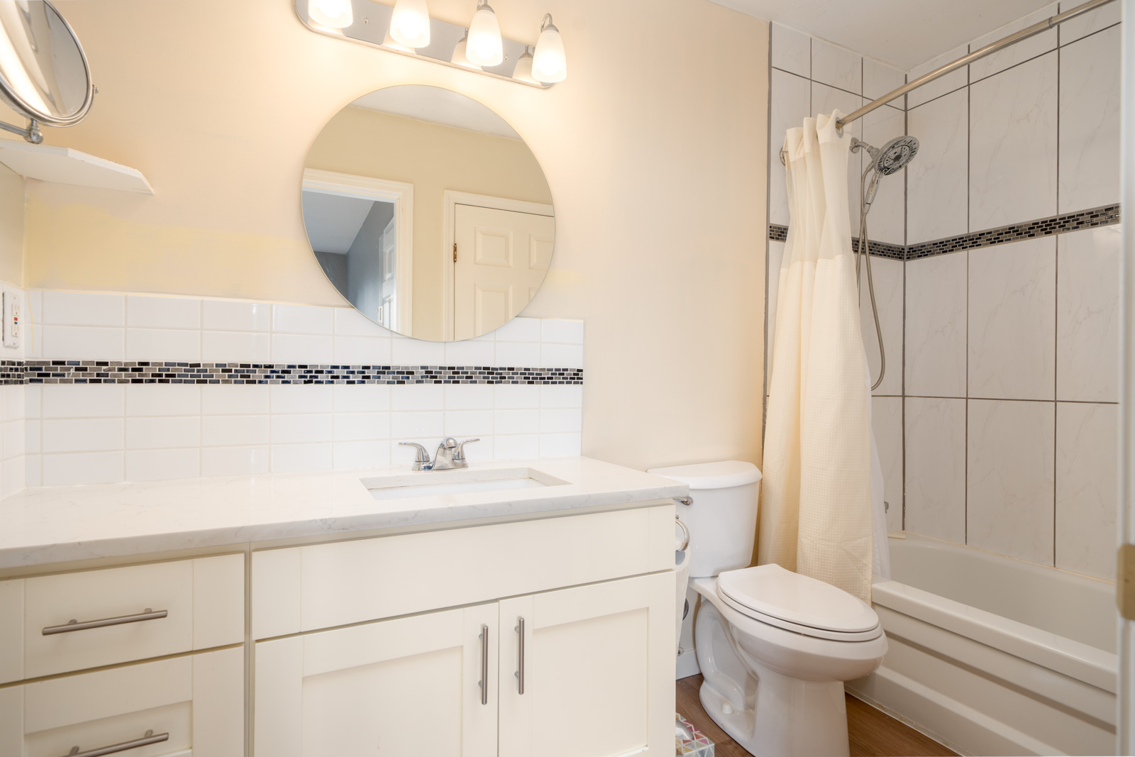 A bathroom with a round mirror above a white vanity, a toilet, and a bathtub with a showerhead and white curtain. The walls have white tile with a black accent strip.