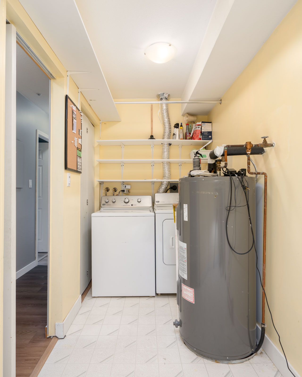Laundry room with a washing machine, dryer, water heater, shelves holding various items, and a corkboard on a yellow wall.