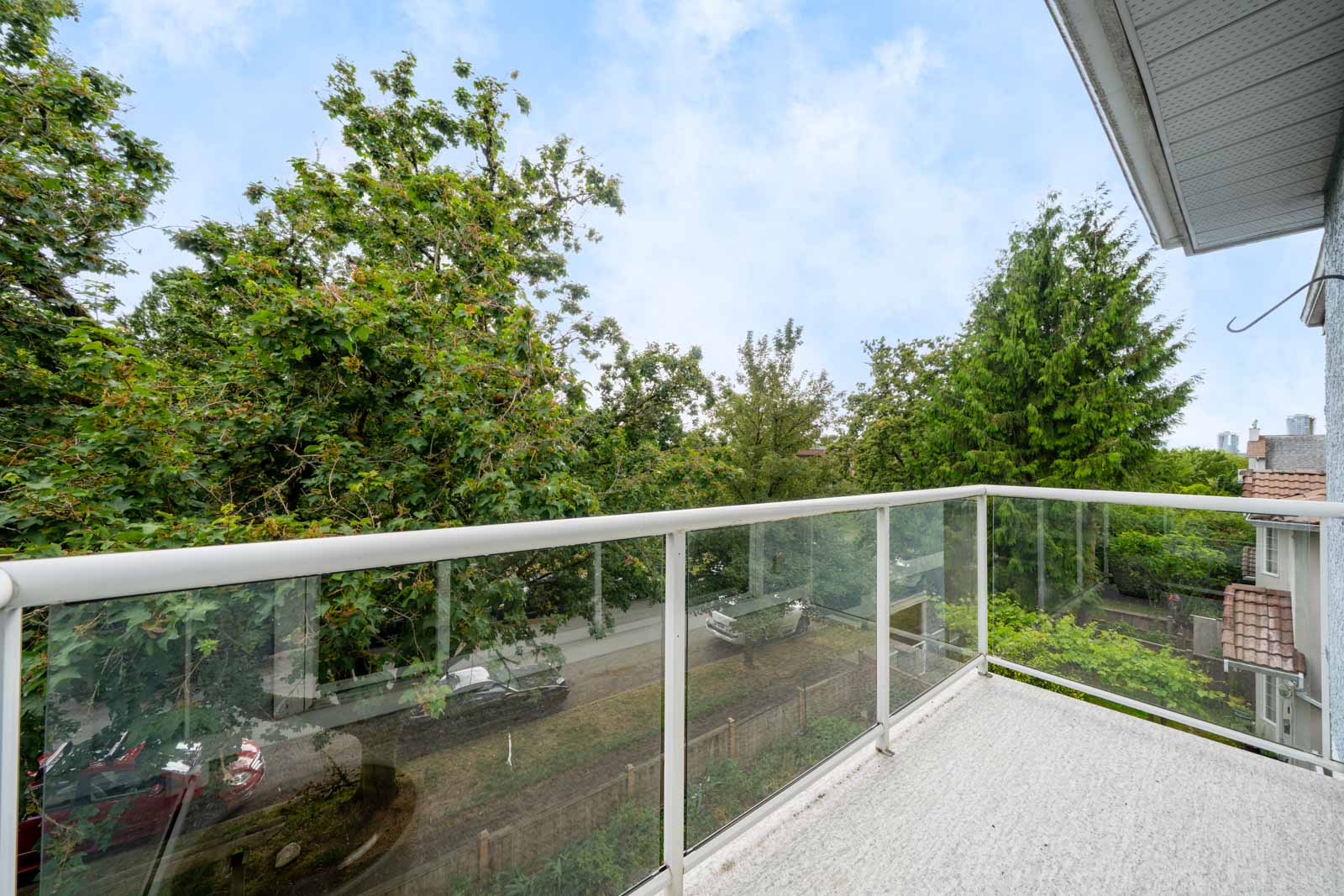 A glass-railed balcony overlooks a street with parked cars, trees, and nearby houses under a partly cloudy sky.