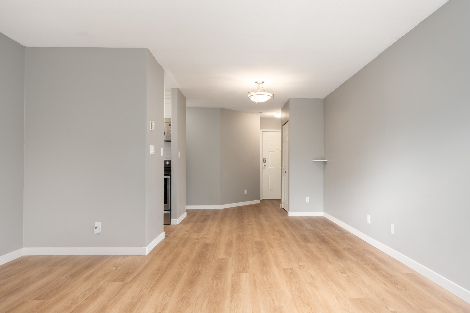 Empty room with light wood flooring, gray walls, white ceiling, a ceiling light fixture, and a doorway leading to the kitchen and entry door.