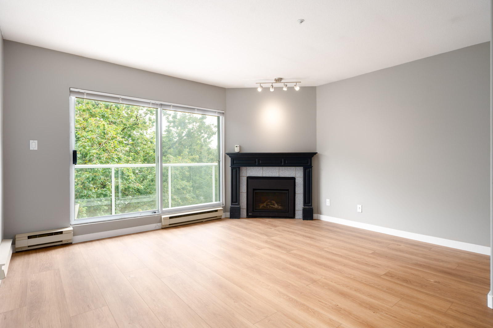 Empty living room with light wood flooring, a black fireplace, large window with sliding glass door, and light gray walls. Green trees are visible outside.