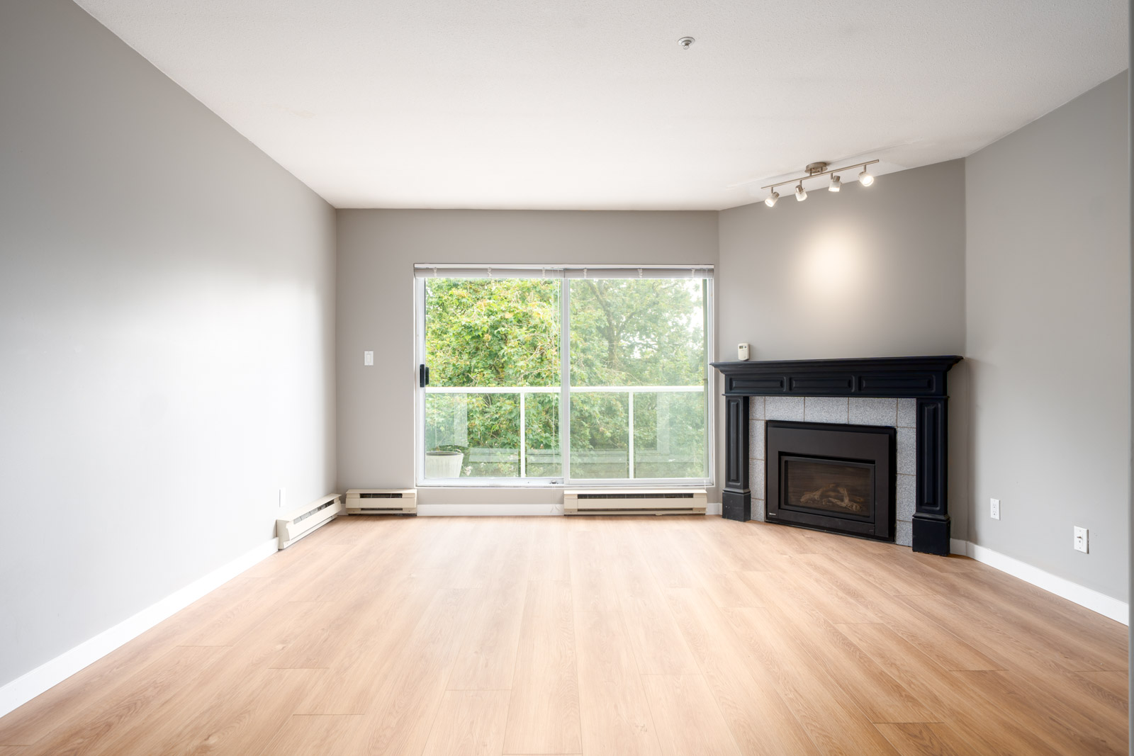 Empty living room with light wood flooring, gray walls, a black fireplace in the corner, large window with a view of trees, and ceiling track lighting.