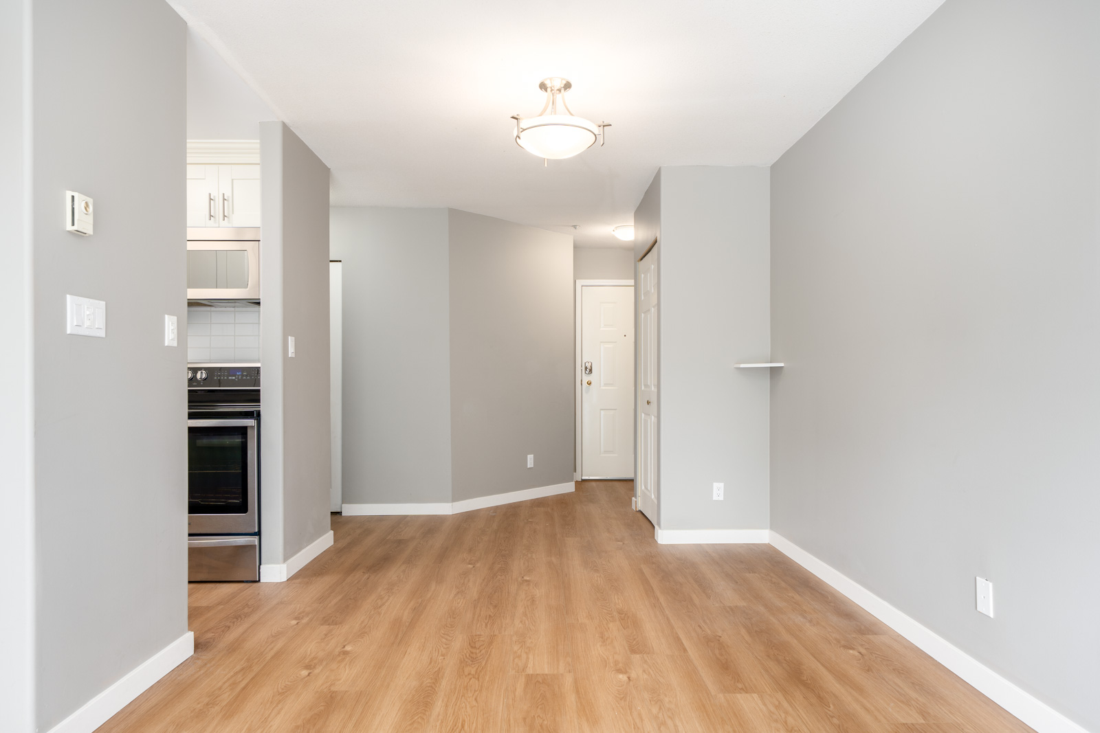 Empty room with light wood flooring, gray walls, white trim, and a ceiling light; kitchen area with stainless steel appliances is partially visible on the left.