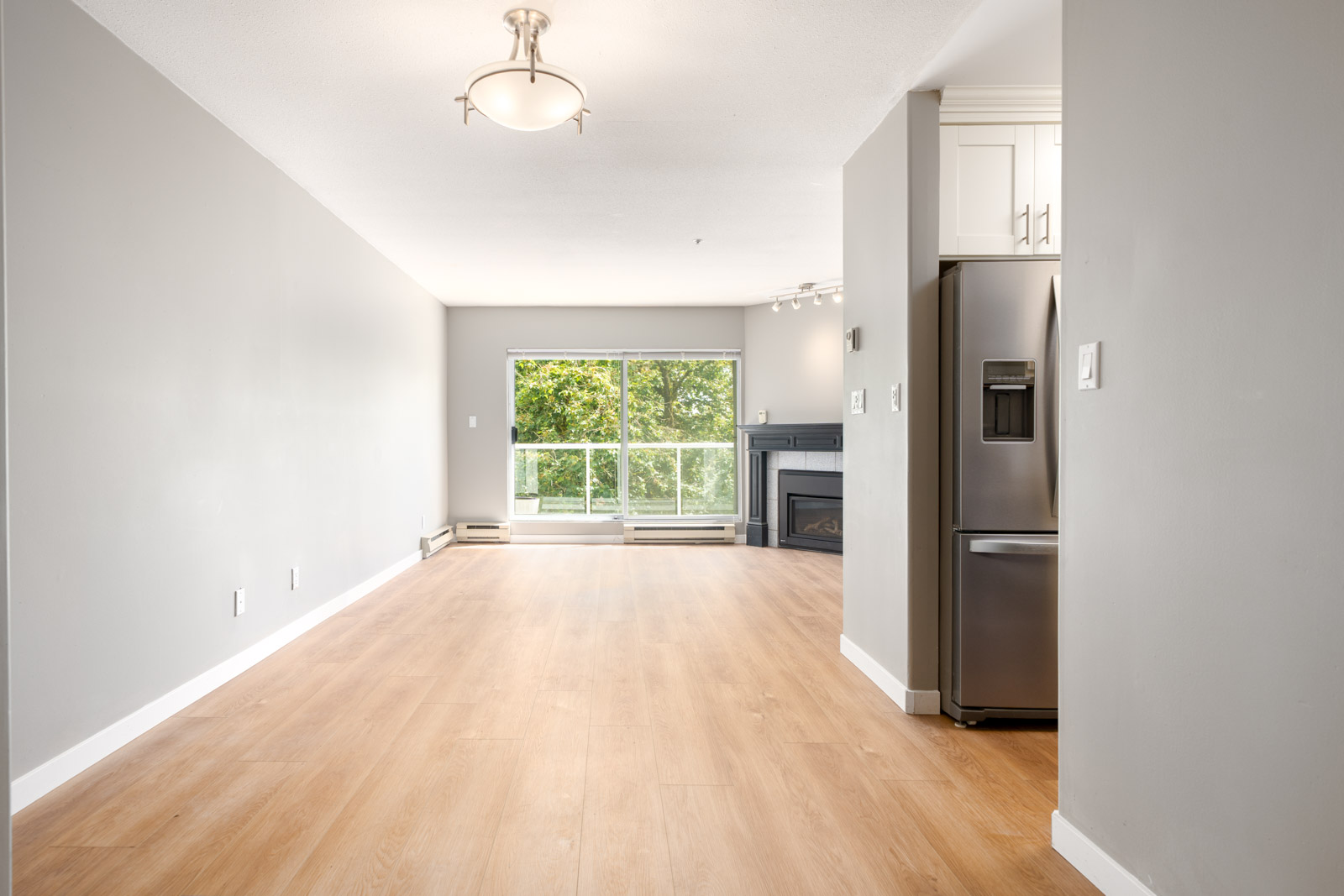 Unfurnished living space with light wood flooring, large window with a view of trees, fireplace, grey walls, and stainless steel refrigerator visible on the right.
