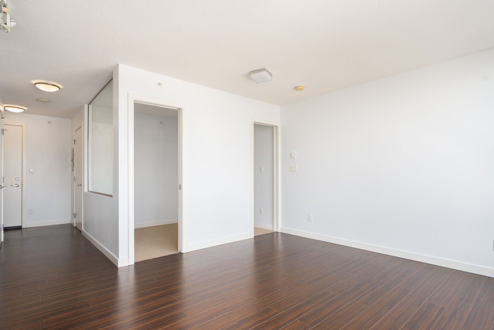 Empty room with white walls, dark wood flooring, two open doorways, a glass partition, and ceiling lights.