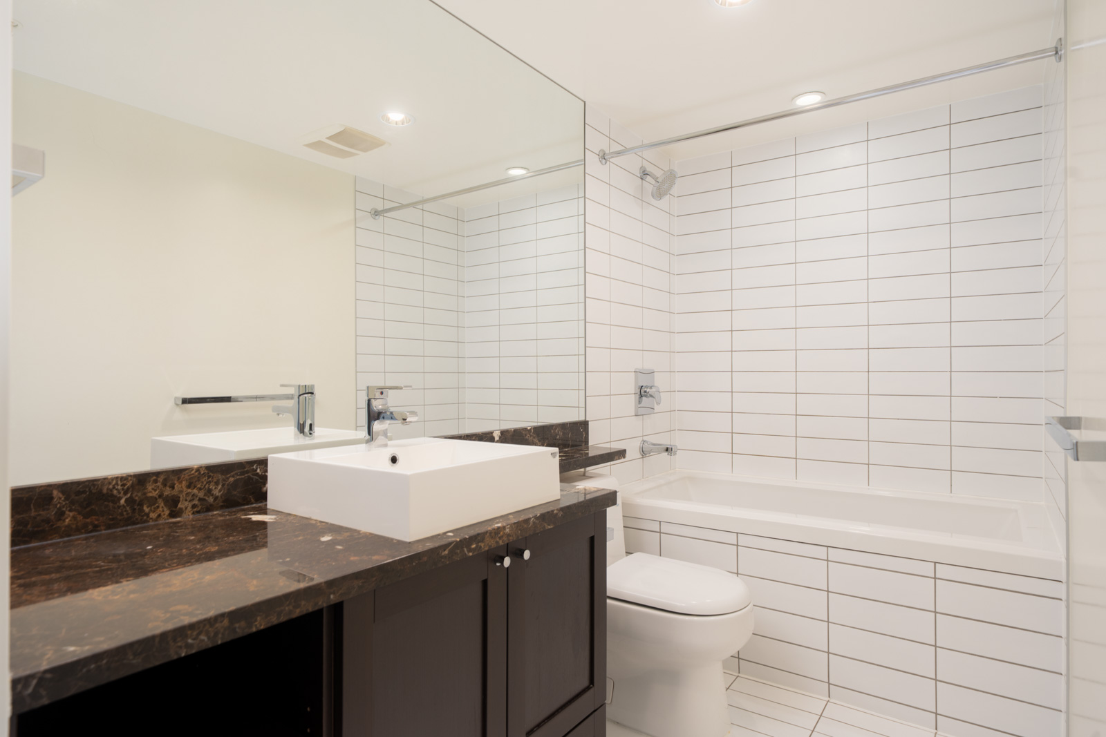 Modern bathroom with a white sink on a dark countertop, large mirror, toilet, and white bathtub with white tile walls and floor.