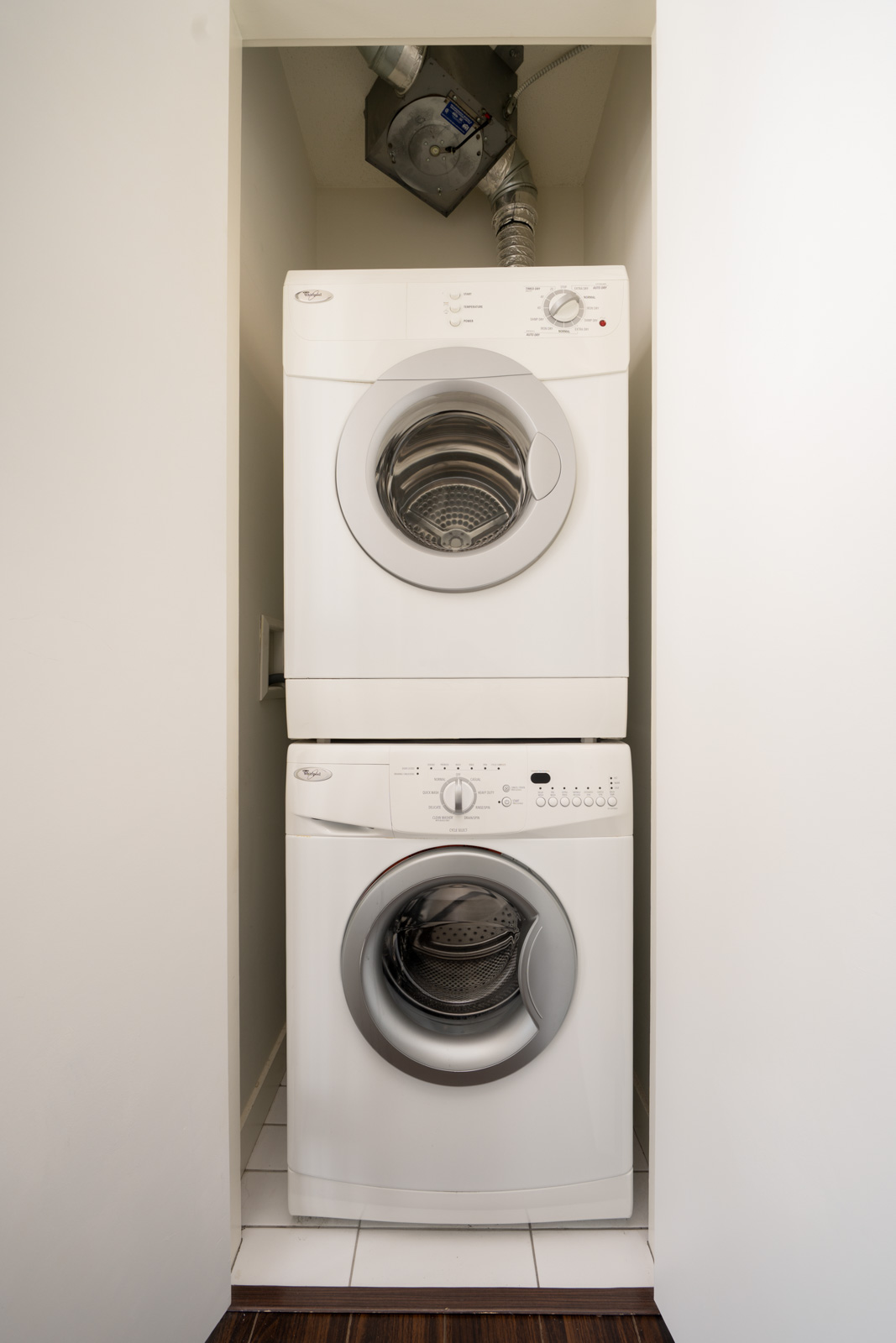 A stacked white washer and dryer are placed in a small, narrow laundry closet with white walls and tile flooring.