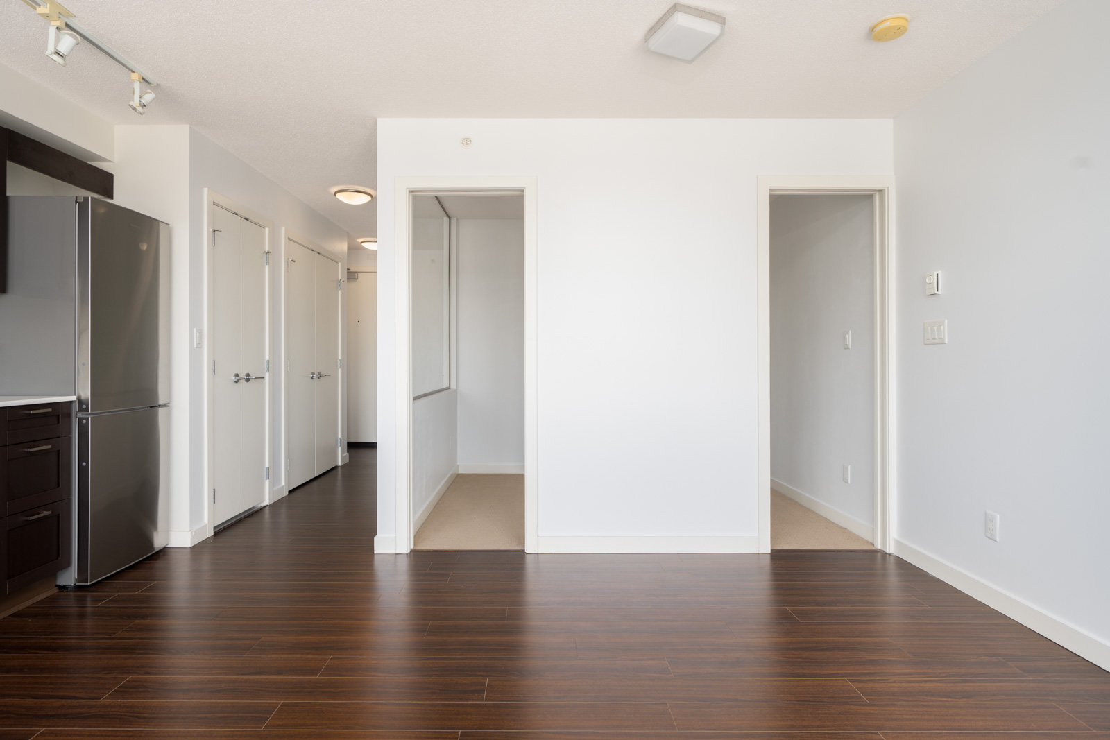 Unfurnished apartment interior with white walls, dark wood flooring, two doorways, a windowed alcove, and a stainless steel refrigerator on the left.