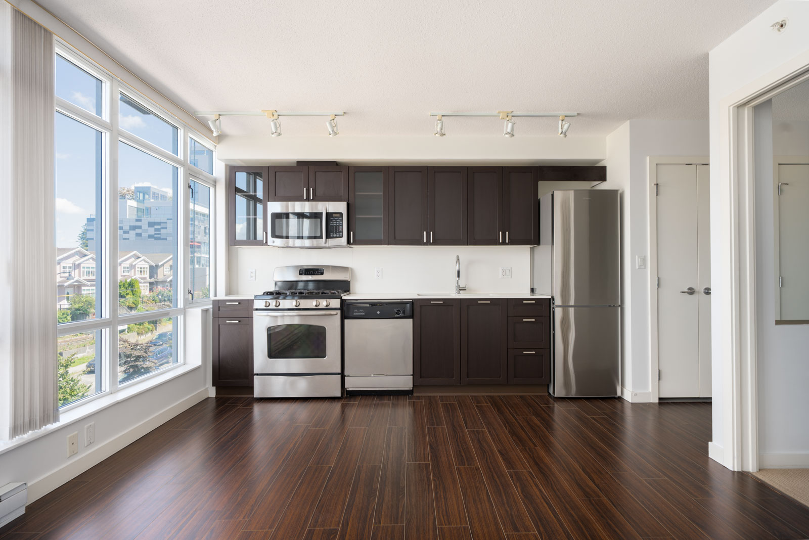 Modern kitchen with dark cabinets, stainless steel appliances, and large windows overlooking a cityscape. The room has hardwood floors and white walls.