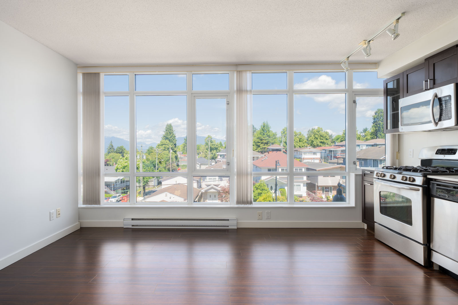 Modern kitchen and dining area with stainless steel appliances, large windows, hardwood floors, and a view of houses and trees outside.