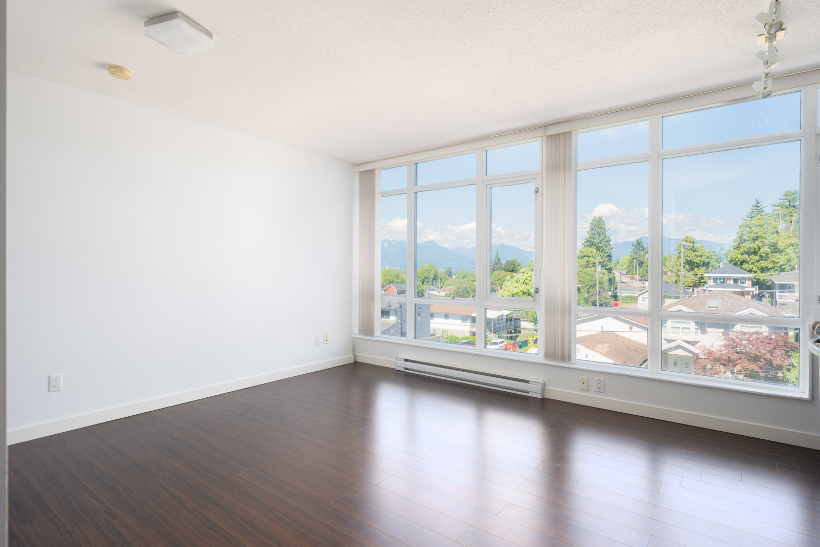 Empty room with white walls, large floor-to-ceiling windows, dark wood flooring, and a view of trees, houses, and mountains outside.