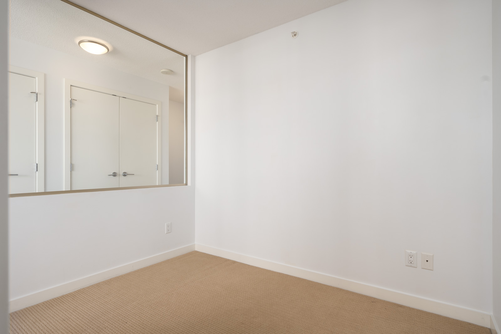 Empty room with beige carpet, white walls, a large mirror, electrical outlets, and a closed double door with overhead light reflected in the mirror.
