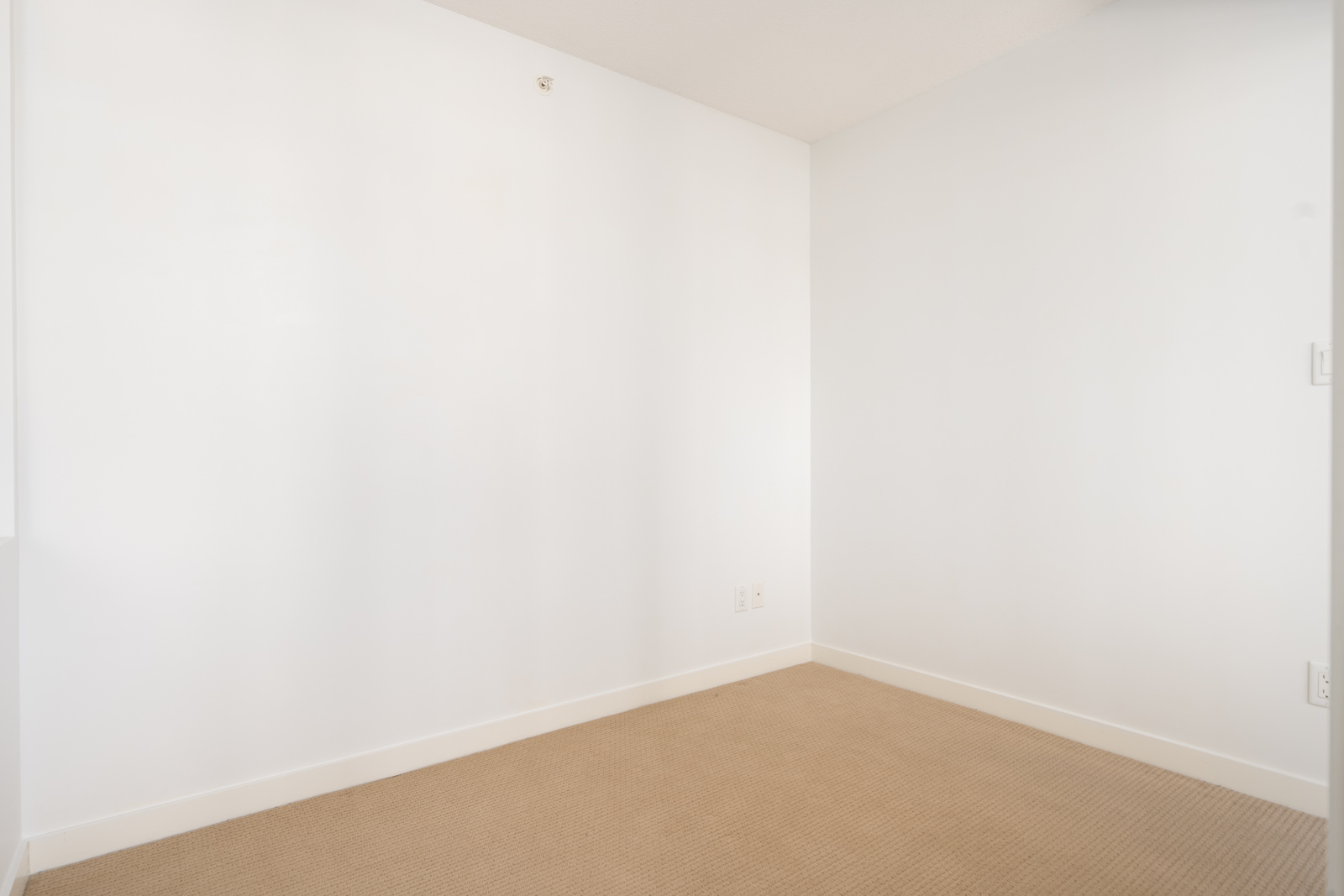 Empty room with white walls and beige carpet flooring, featuring electrical outlets and no furniture or decorations.