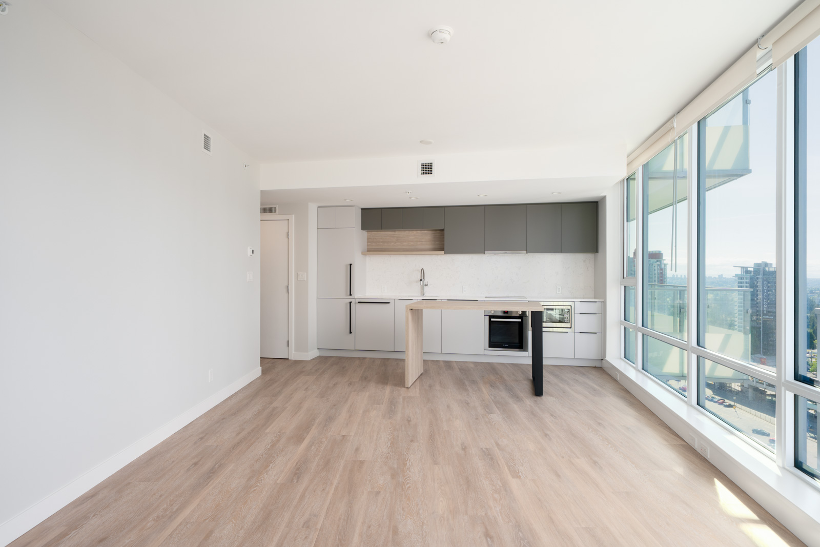 Modern, unfurnished apartment with light wood floors, white walls, built-in kitchen, and large floor-to-ceiling windows offering a city view.