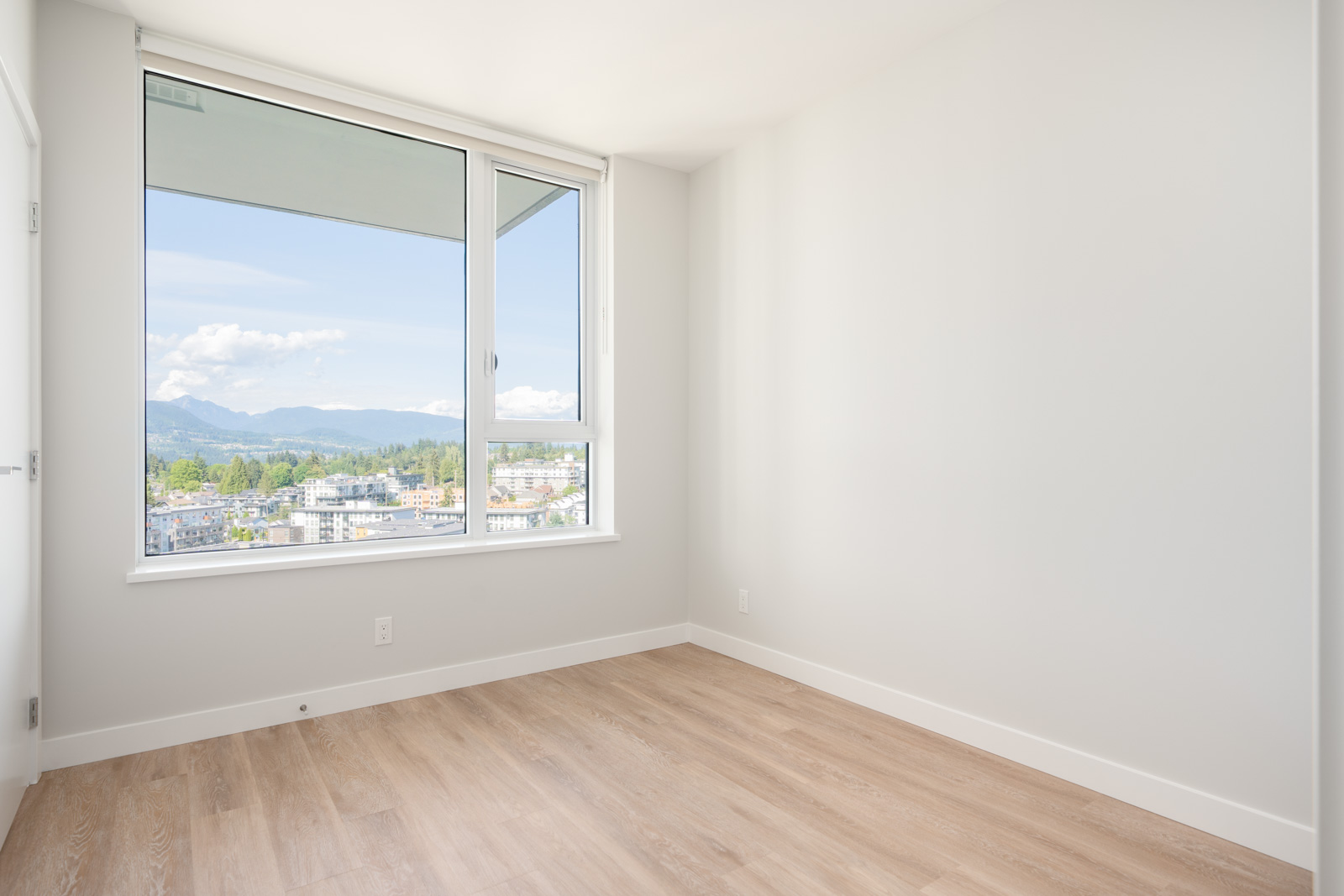 Empty room with light wood flooring, white walls, and a large window showing a cityscape and mountains under a blue sky.
