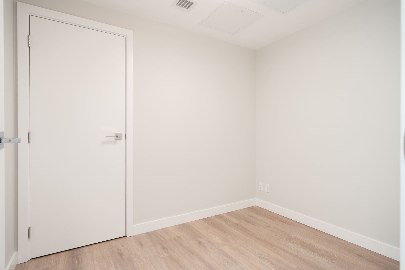 Empty, unfurnished room with white walls, a closed white door, light wood flooring, and two visible electrical outlets in the corner.