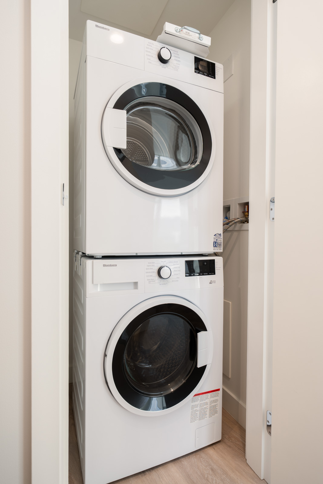 A front-loading washer and dryer are stacked vertically in a small laundry closet with white walls and wood flooring.
