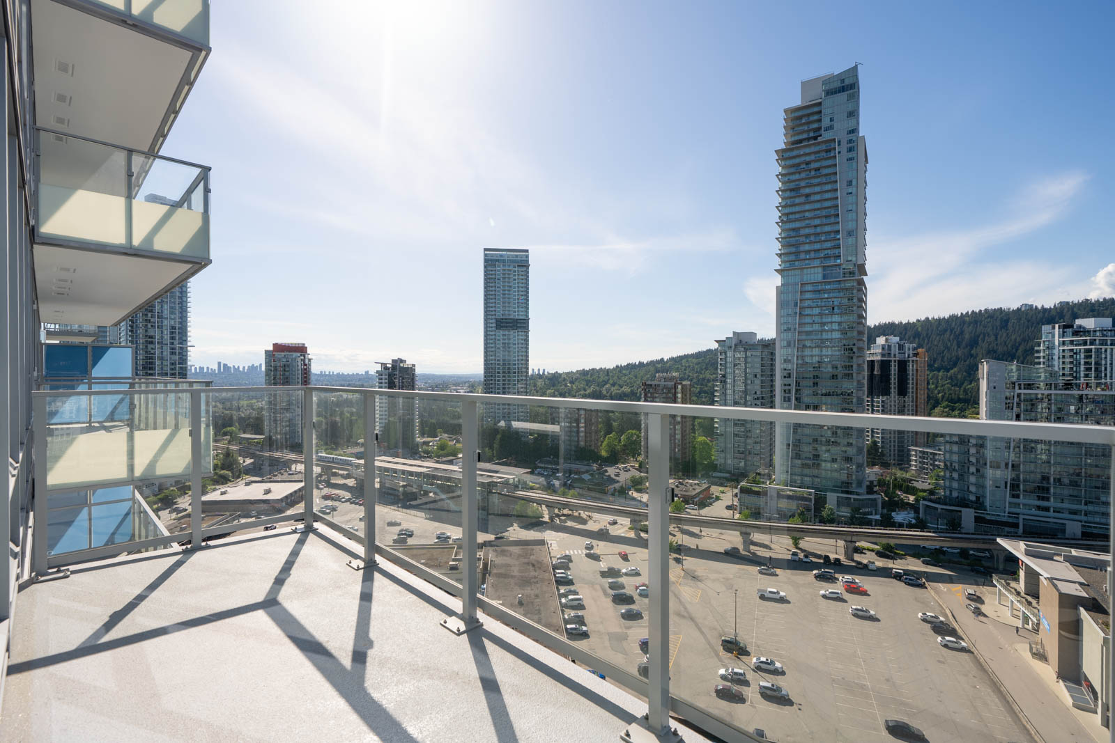 View from a high-rise balcony overlooking a busy street, parking lot, and several tall buildings under a clear blue sky.