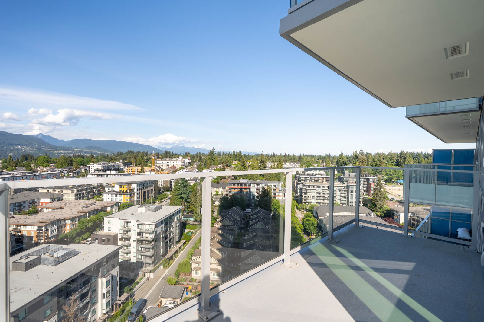 High-rise balcony with glass railing overlooking a cityscape of mid-rise buildings, trees, and distant mountains under a clear blue sky.